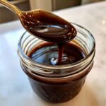 A spoonful of thick, glossy homemade chocolate syrup drips back into a small glass jar.