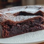 Close-up of a rich, dark flourless chocolate torte with a slice cut out, revealing a fudgy interior, dusted with powdered sugar.