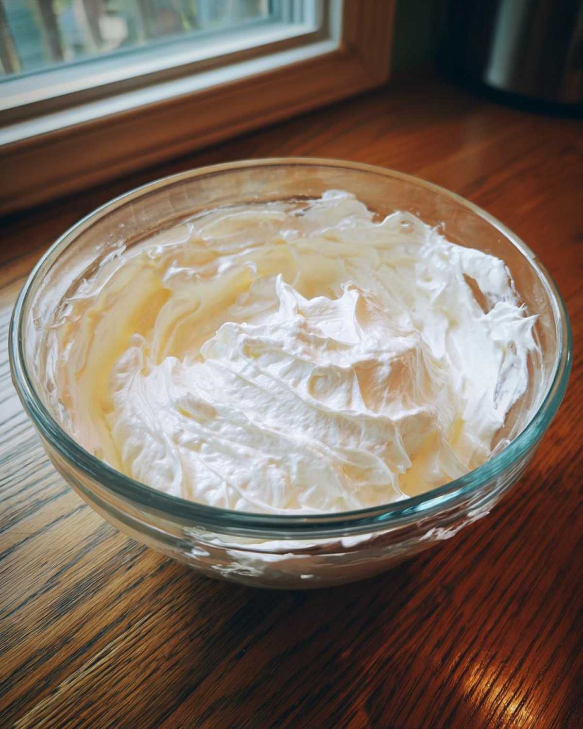Close-up of thick, white, fluffy royal icing recipe mixture in a clear glass bowl on a wooden surface.