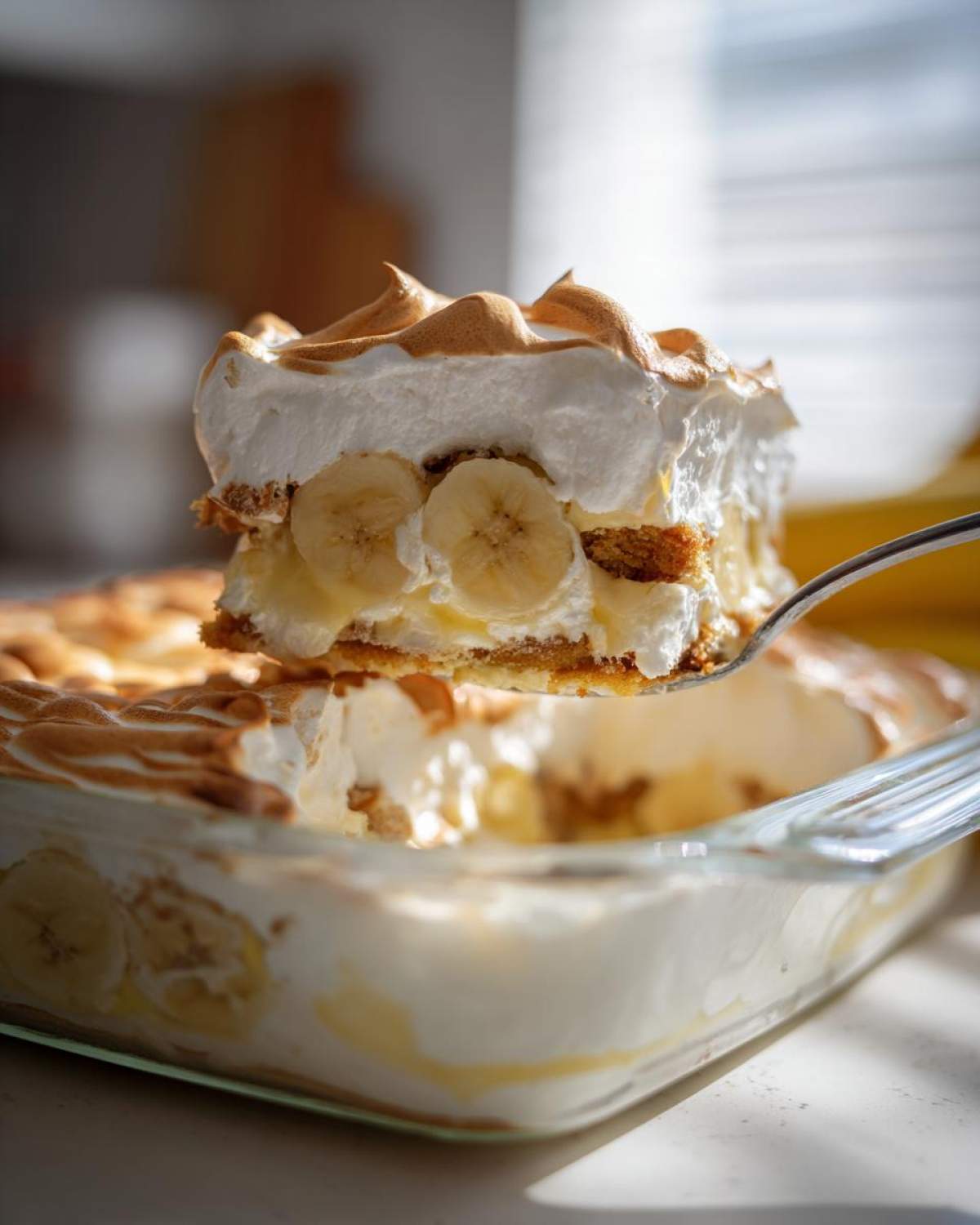 A spoonful of layered banana pudding with meringue topping being lifted from a glass dish.