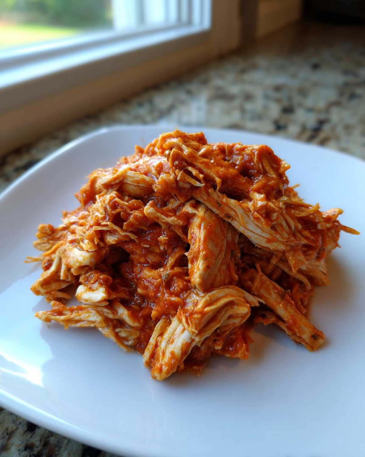 A close-up of shredded chicken tinga coated in a rich, orange-red sauce, served on a square white plate.