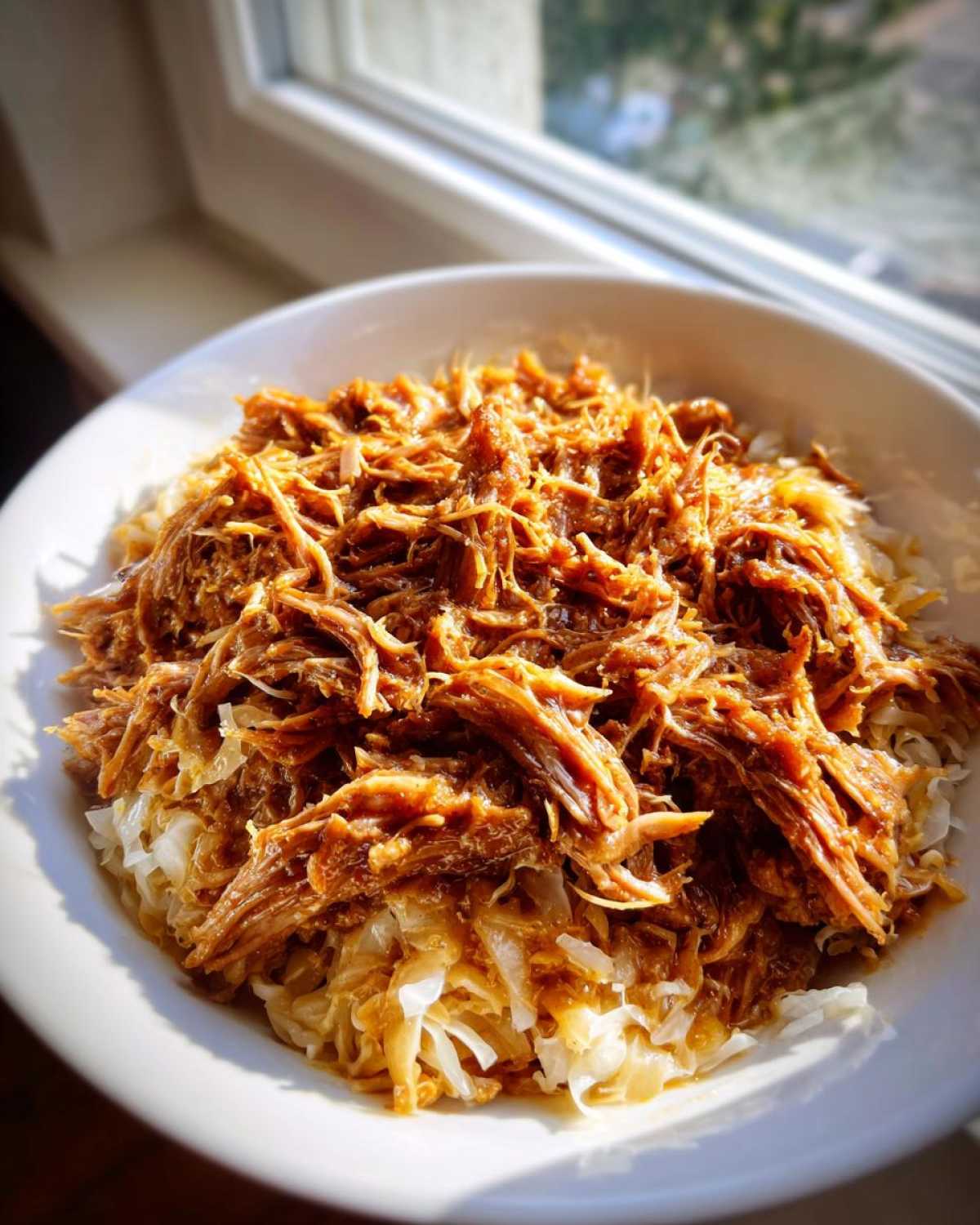 Close-up of shredded pork piled high over a bed of sauerkraut in a white bowl, backlit by a window.