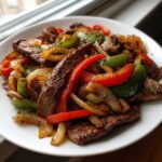 Close-up of sizzling steak fajitas featuring seasoned steak strips, sautéed red and green bell peppers, and onions on a white plate.