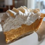 A close-up slice of rich, orange-hued butterscotch pie topped generously with piped white whipped cream.