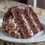 A decadent slice of german chocolate cake recipe showing moist dark layers, white filling, and toasted coconut flakes on the side.