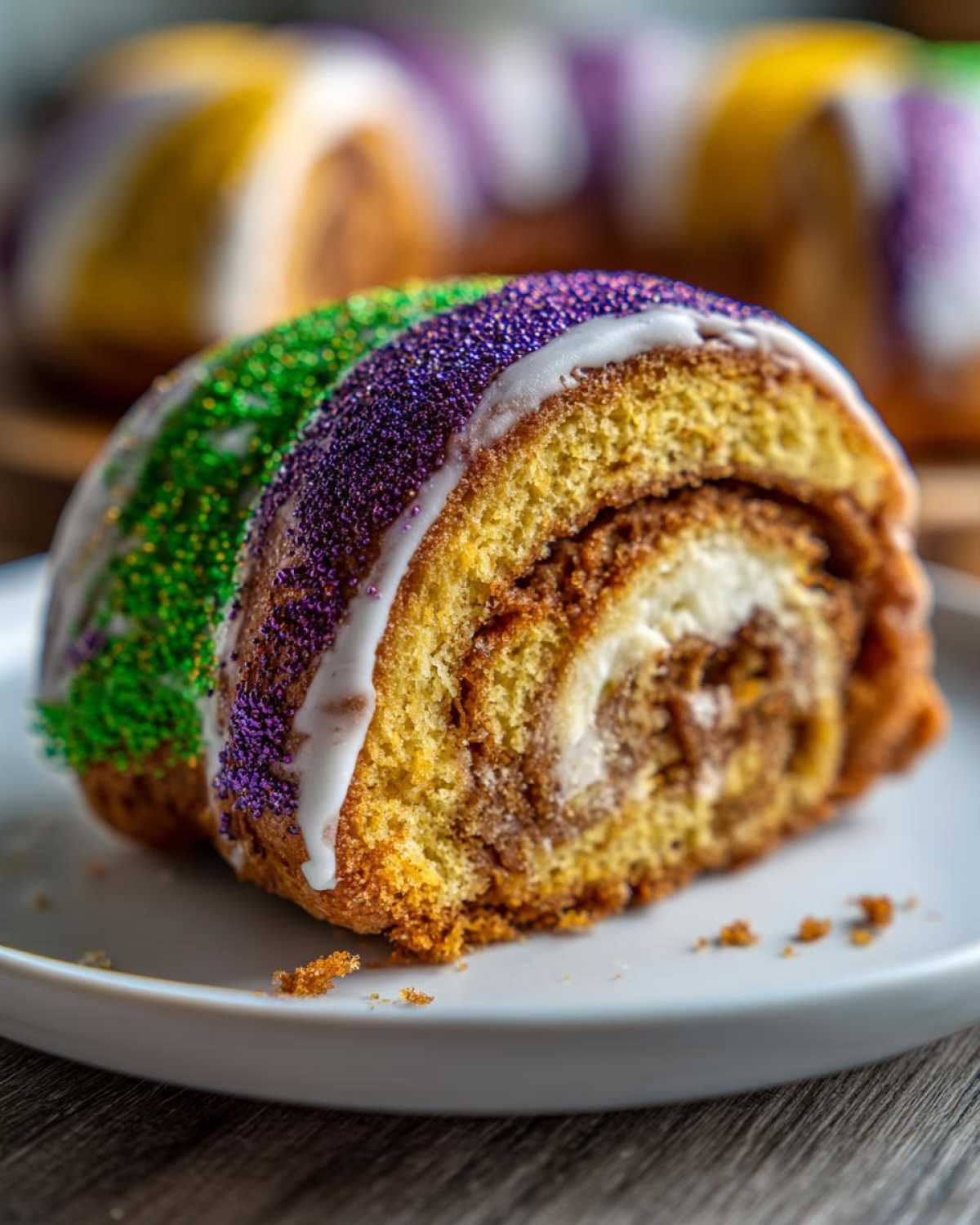 Close-up of a slice of cream cheese king cake, showing the swirl filling and topped with white icing and purple and green Mardi Gras sprinkles.