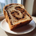Close-up of a thick slice of homemade raisin bread with visible swirls of dark raisins on a white plate.