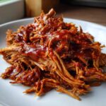 Close-up of shredded, saucy Slow cooker pulled pork piled high on a white plate.