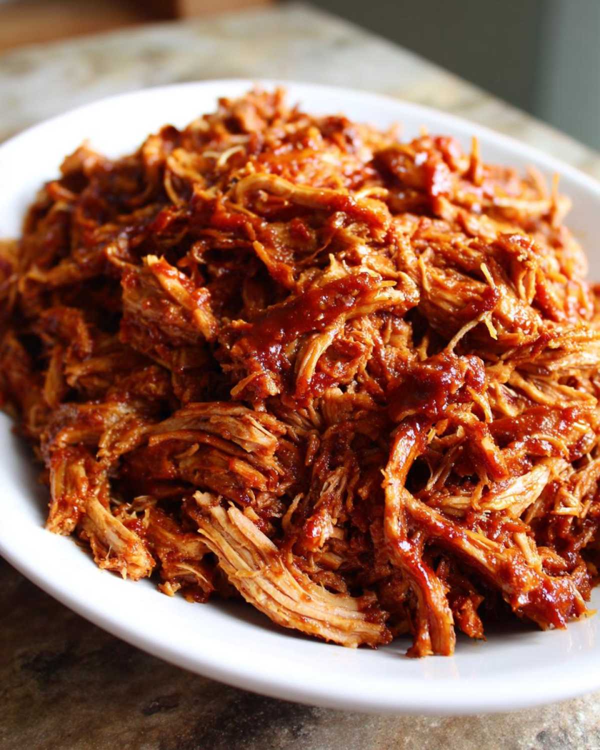 A close-up shot of shredded, saucy Slow cooker pulled pork piled high on a white oval serving platter.