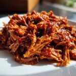 Close-up of tender, shredded Slow cooker pulled pork coated in rich barbecue sauce, served on a white plate.