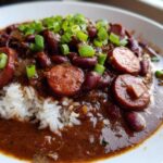 A close-up of a bowl of rich, dark slow cooker red beans and rice topped with sliced smoked sausage and green onions.