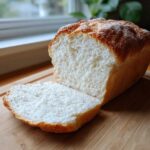 A loaf of freshly baked, soft condensed milk bread with one thick slice cut and resting beside it on a wooden board.