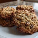 Four freshly baked, soft oatmeal cookie treats stacked slightly on a white plate, featuring visible oats and chocolate chips.