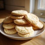 A generous stack of freshly baked soft sugar cookies, lightly coated in sugar, sitting on a light plate.