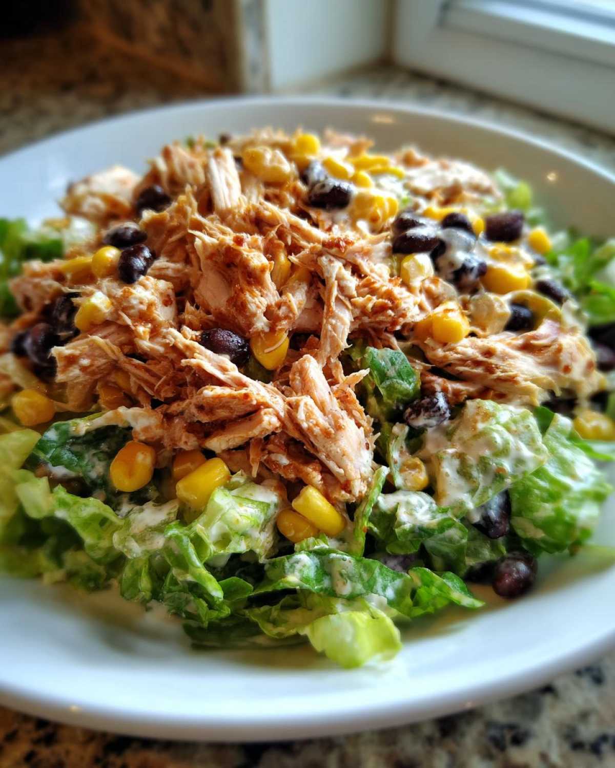 Close-up of a bowl of southwest chicken salad featuring shredded seasoned chicken, black beans, corn, and creamy dressing over lettuce.