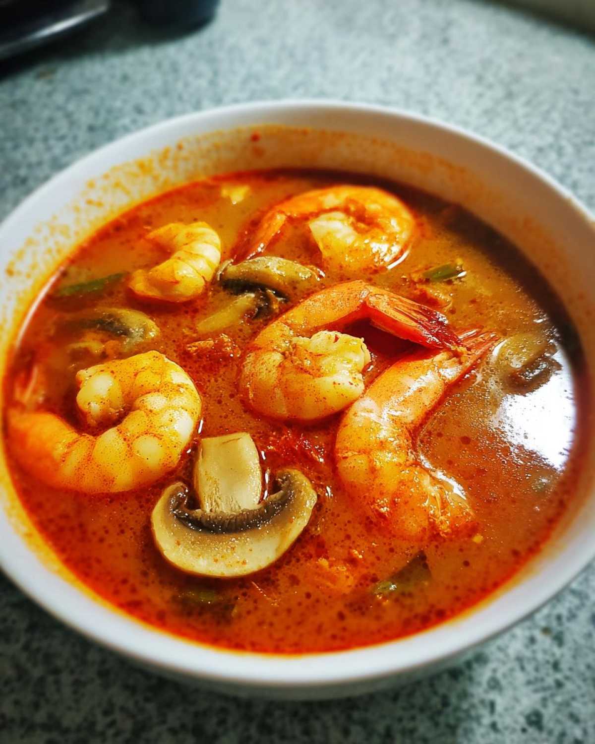 Close-up of a white bowl filled with vibrant red-orange tom yum soup, featuring large shrimp and sliced mushrooms.