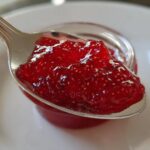 Close-up of a spoonful of glistening, bright red homemade raspberry jam showing visible seeds.
