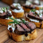 Close-up of perfectly cooked steak crostini appetizers topped with cheese, balsamic glaze, and herbs on a wooden cutting board.