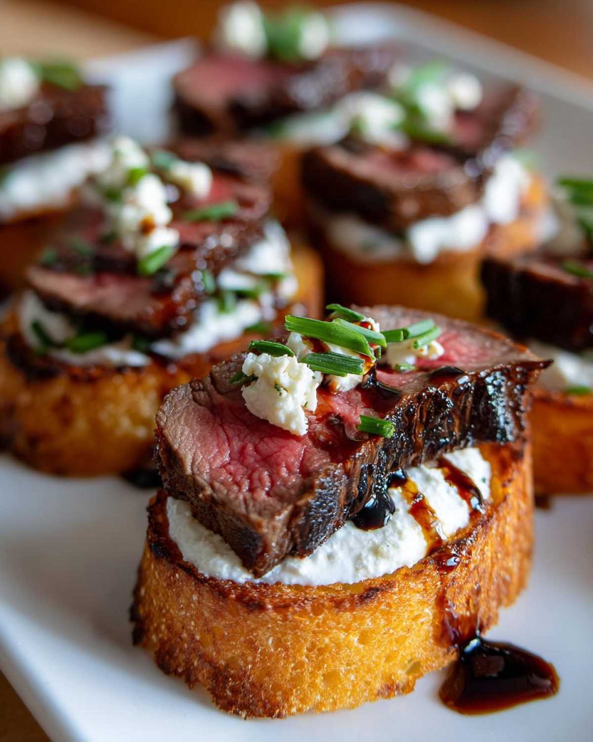 Close-up of a single steak crostini topped with medium-rare steak, cheese, and chives, drizzled with balsamic glaze.