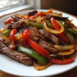 A close-up of vibrant steak fajitas featuring sliced steak, red and green bell peppers, and onions on a white plate.