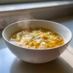A steaming white bowl filled with golden yellow egg drop soup, showing wisps of steam rising.