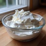 A glass bowl filled with thick, stiff white royal icing recipe mixture sitting on a wooden countertop near a window.