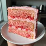 Close-up of a moist slice of two-layer strawberry cake with pink frosting and visible strawberry pieces.