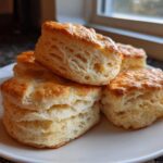 Close-up of several tall, golden-brown buttermilk biscuits showing flaky layers, stacked on a white plate.