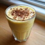 A tall glass filled with thick, creamy yellow eggnog, topped with a dusting of cinnamon or nutmeg.