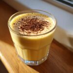 A glass of thick, creamy yellow eggnog topped with a generous dusting of ground nutmeg.