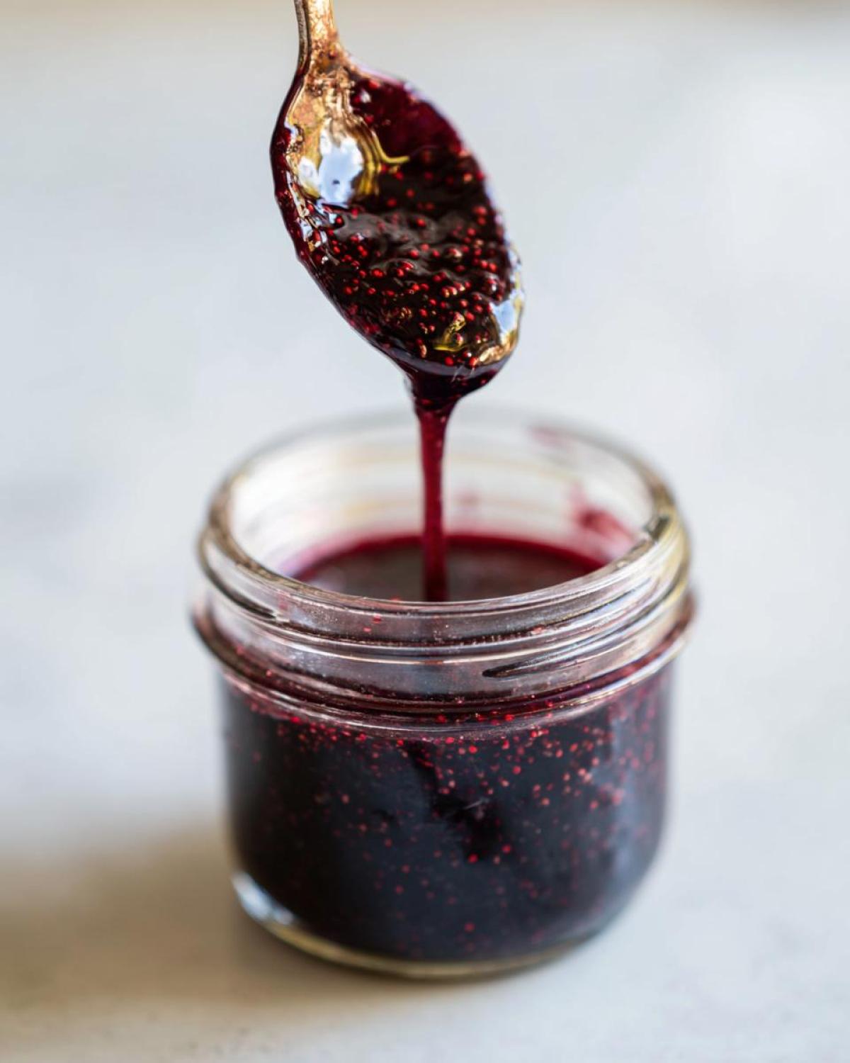 A spoonful of thick, dark elderberry syrup, showing seeds, dripping back into a small glass jar.