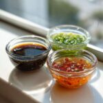 Three small glass bowls containing different types of Dumpling Sauce: dark soy-based, chili oil, and green scallion sauce.