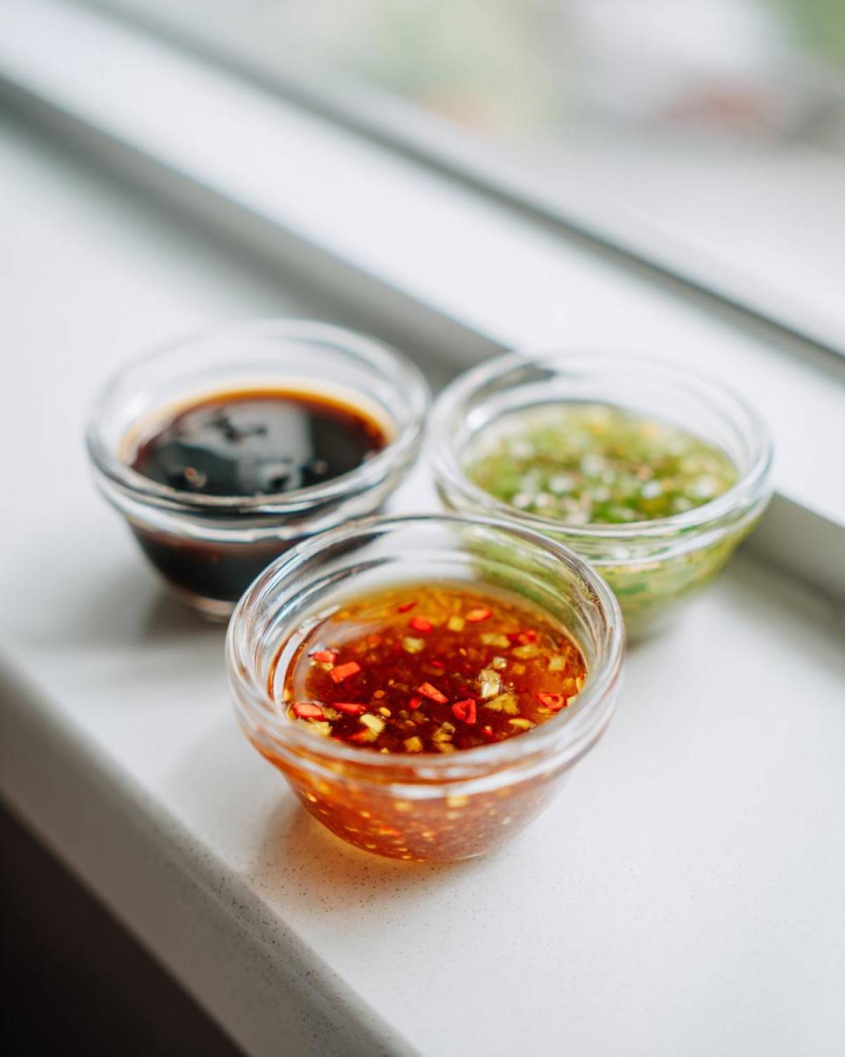 Three small glass bowls containing different types of dumpling sauce, including a chili sauce, a green sauce, and a dark soy-based sauce.