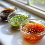 Three small glass bowls containing different types of Dumpling Sauce, including a dark soy-based sauce, a green scallion sauce, and a chili garlic sauce.