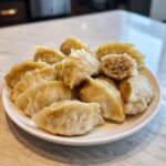 A white plate piled high with freshly steamed Traditional Chinese Dumplings, one cut open to show the savory filling.