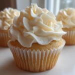 Close-up of a beautifully frosted vanilla cupcake with rich buttercream swirls, two more vanilla cupcakes blurred in the background.