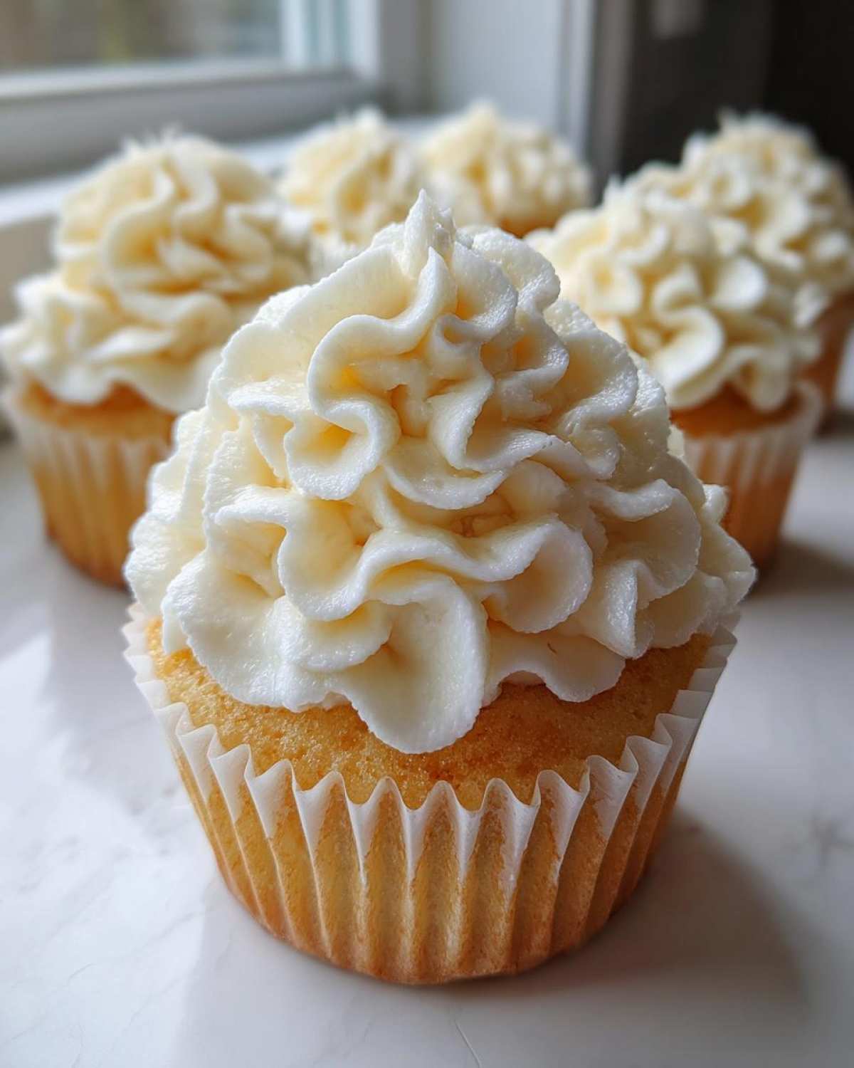 A close-up of a freshly baked vanilla cupcake topped with a generous swirl of creamy white frosting, with more vanilla cupcakes blurred in the background.