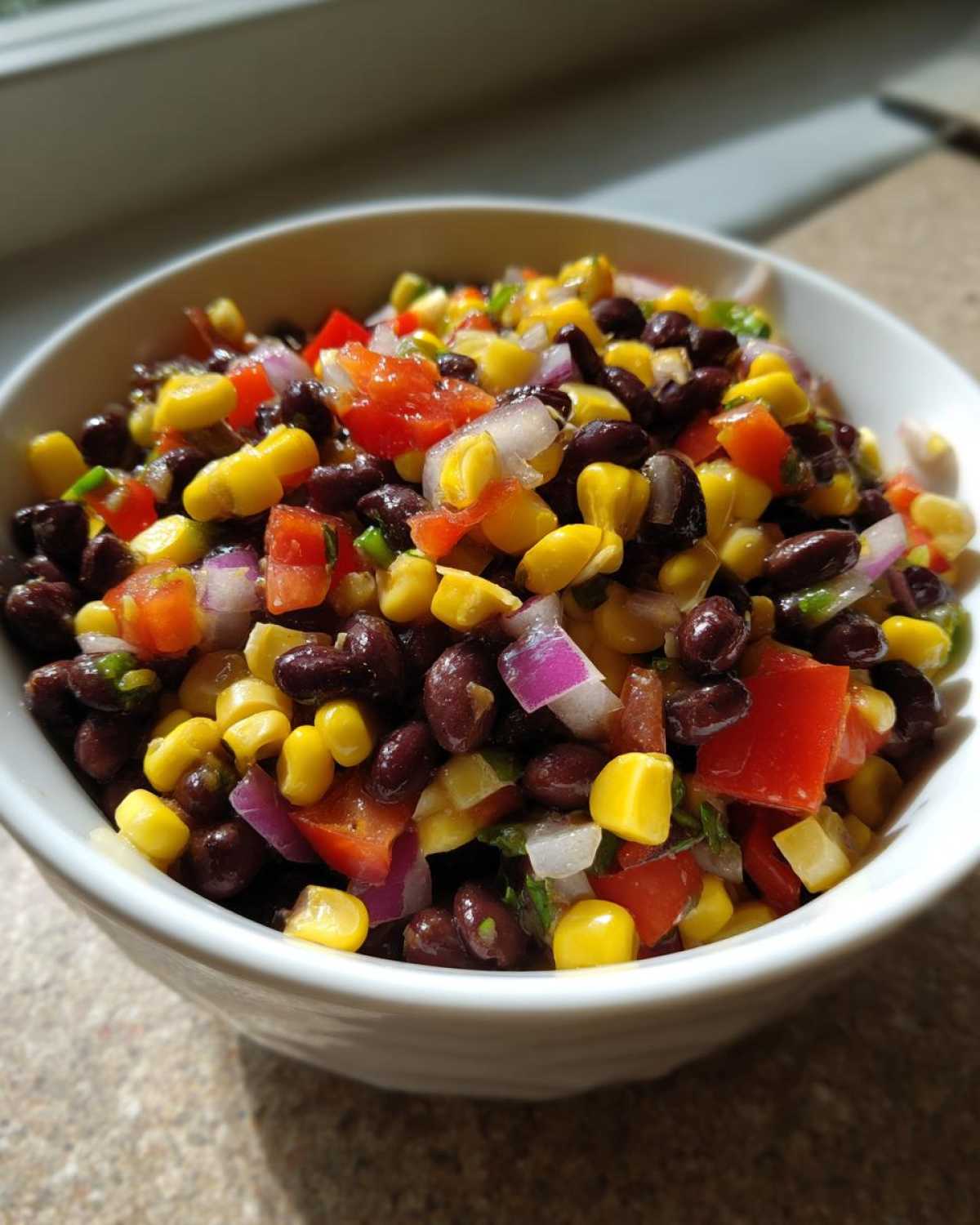 Close-up of a white bowl filled with colorful cowboy caviar, featuring black beans, corn, red onion, and tomato.