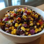 Close-up of a white bowl filled with colorful cowboy caviar featuring black beans, corn, red onion, and tomatoes.