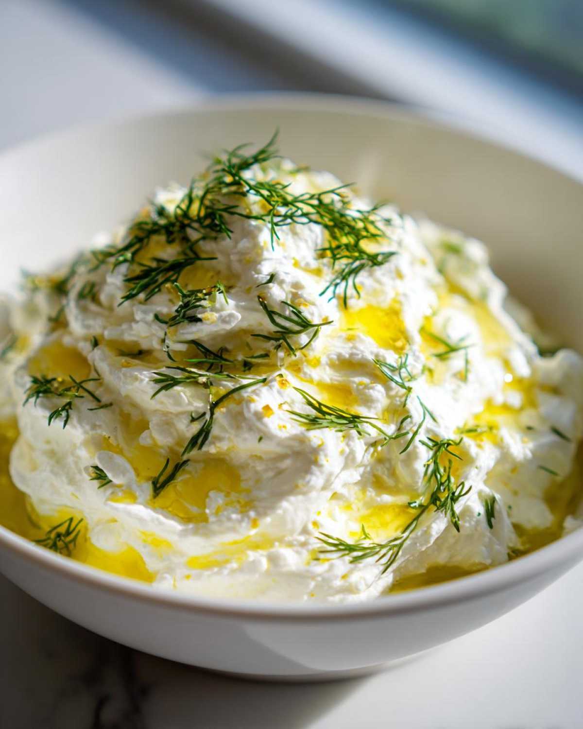 Close-up of creamy whipped feta dip in a white bowl, drizzled with olive oil and fresh dill.