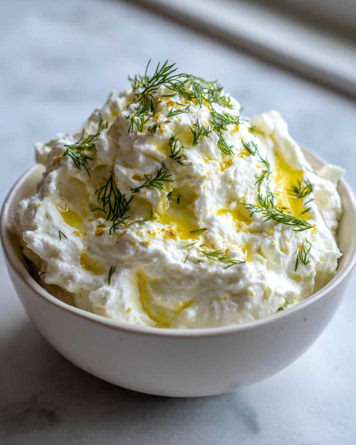 A close-up of creamy whipped feta dip garnished with fresh dill and a drizzle of olive oil.