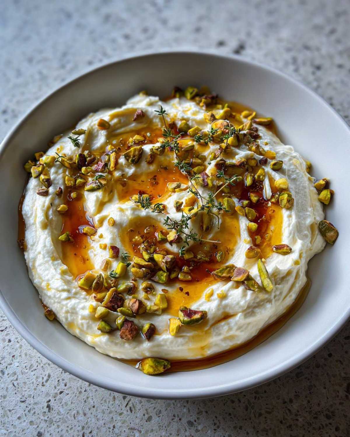 A bowl of creamy whipped goat cheese topped with honey, chopped pistachios, and fresh thyme sprigs.