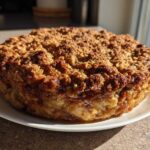 A whole, freshly baked apple crisp recipe dessert with a thick, crunchy topping sitting on a white plate.