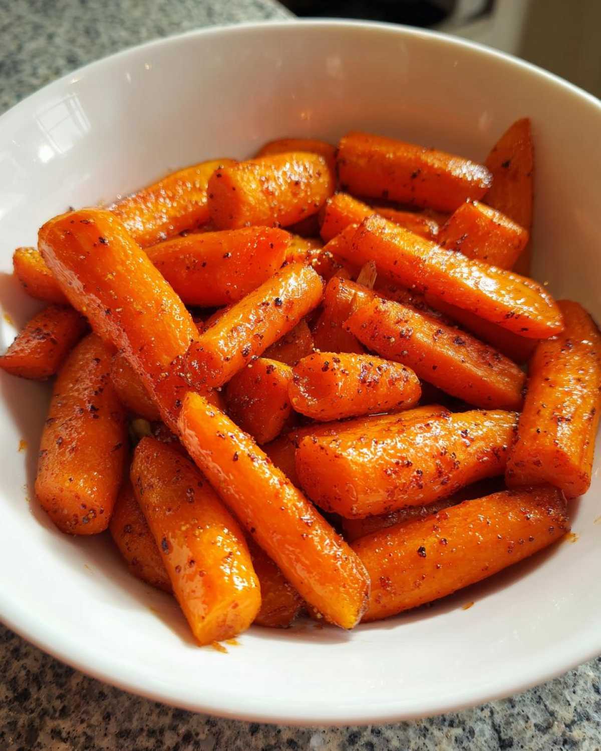 A white bowl filled with shiny, glazed Air Fryer Maple Glazed Carrots sprinkled with spices.