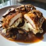 Cross-section of breaded Copycat Olive Garden Stuffed Chicken Marsala showing melted cheese and ham, topped with mushrooms and brown sauce.