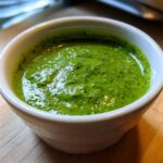 Close-up of bright green Copycat Panera Green Goddess Dressing in a small white bowl on a wooden surface.