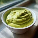 A close-up of thick, bright green Creamy Avocado Cilantro Sauce swirled in a white bowl near a window.