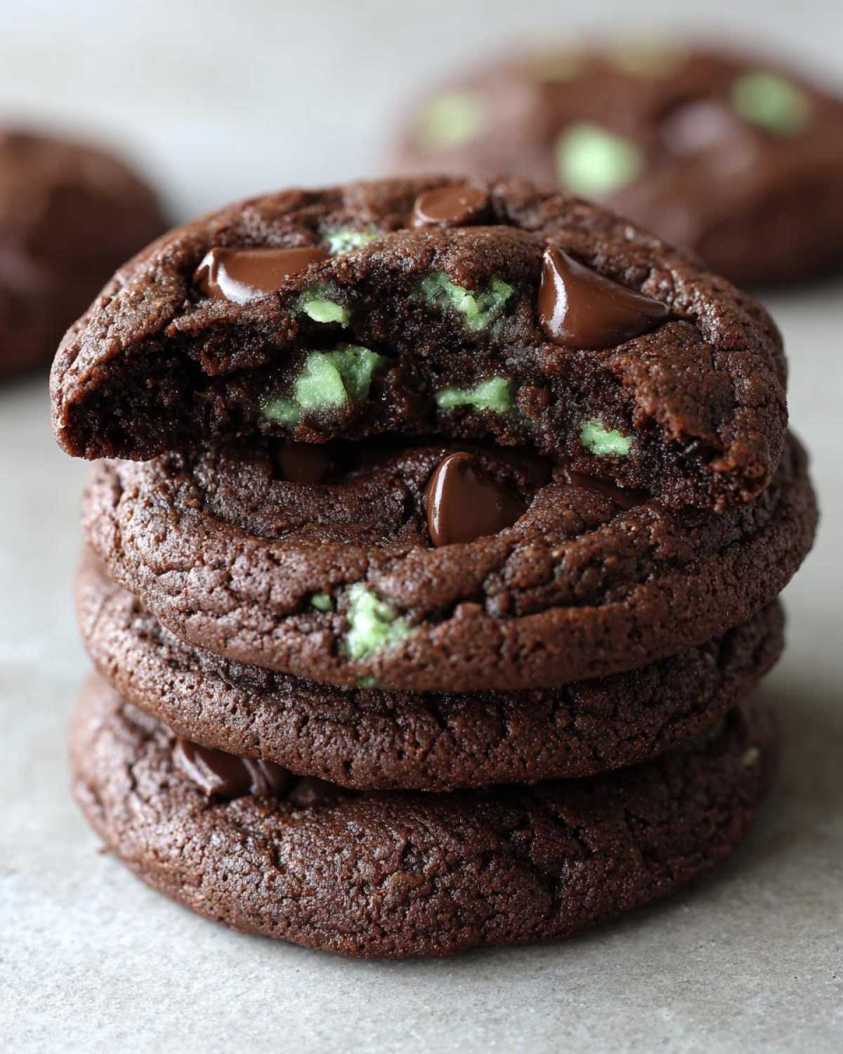 Close-up of a stack of three rich, dark chocolate cookies, with the top one broken open to show gooey chocolate chips and bright green mint chips in these Gluten-Free Chocolate Mint Cookies.