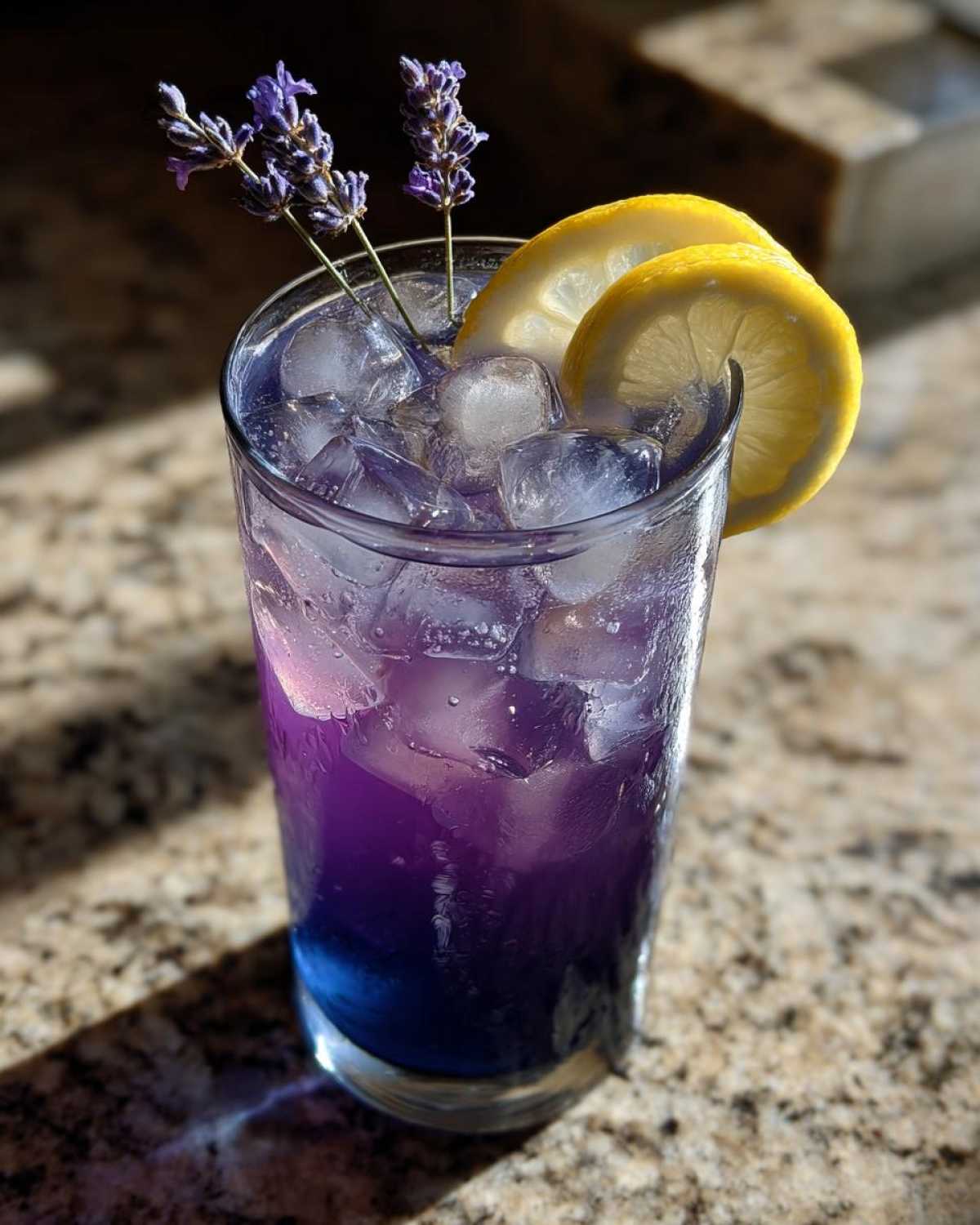 Close-up of a tall glass filled with Iced Blueberry Lavender Lemonade, ice, lemon slices, and lavender sprigs.