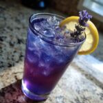 A tall glass filled with vibrant purple and blue Iced Blueberry Lavender Lemonade, ice, and garnished with a lemon slice and lavender sprig.